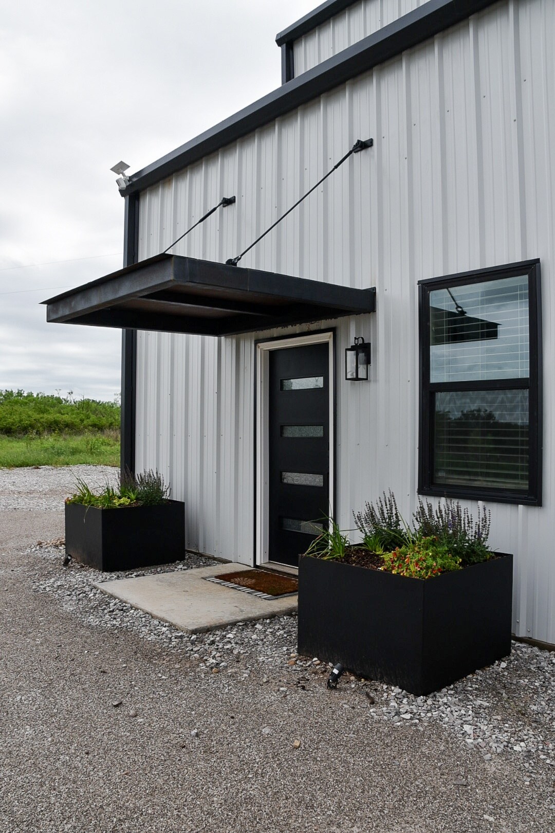 The entrance features a modern black door with sleek horizontal panels, complemented by a contemporary wall light. Flanking the entrance are two large planter boxes filled with greenery. A concrete path leads to the door, surrounded by gravel.