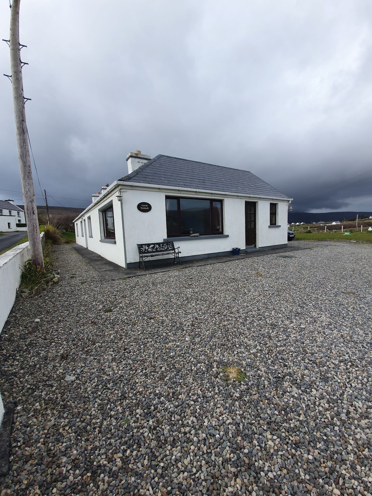 A single-story cottage is displayed, featuring a slate roof and white exterior. A large window provides views of the surroundings. The gravel driveway is lined with grass, and a bench is positioned near the entrance, inviting visitors to relax outdoors.