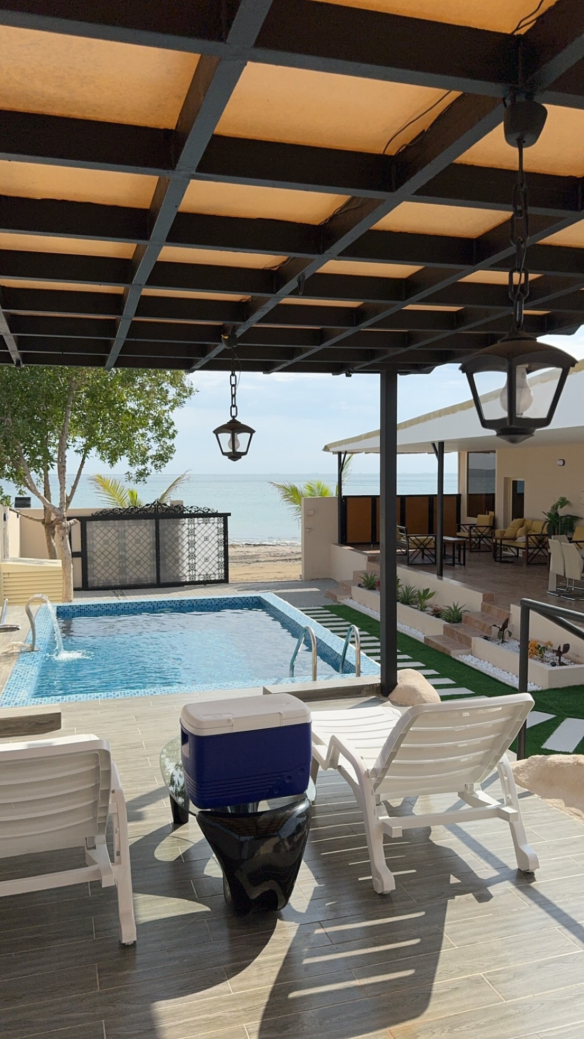 A serene outdoor area features a sparkling pool surrounded by lounge chairs, with a view of sandy beach and calm waters beyond. The space is shaded by a pergola, and decorative elements, including lanterns and greenery, enhance the coastal setting.