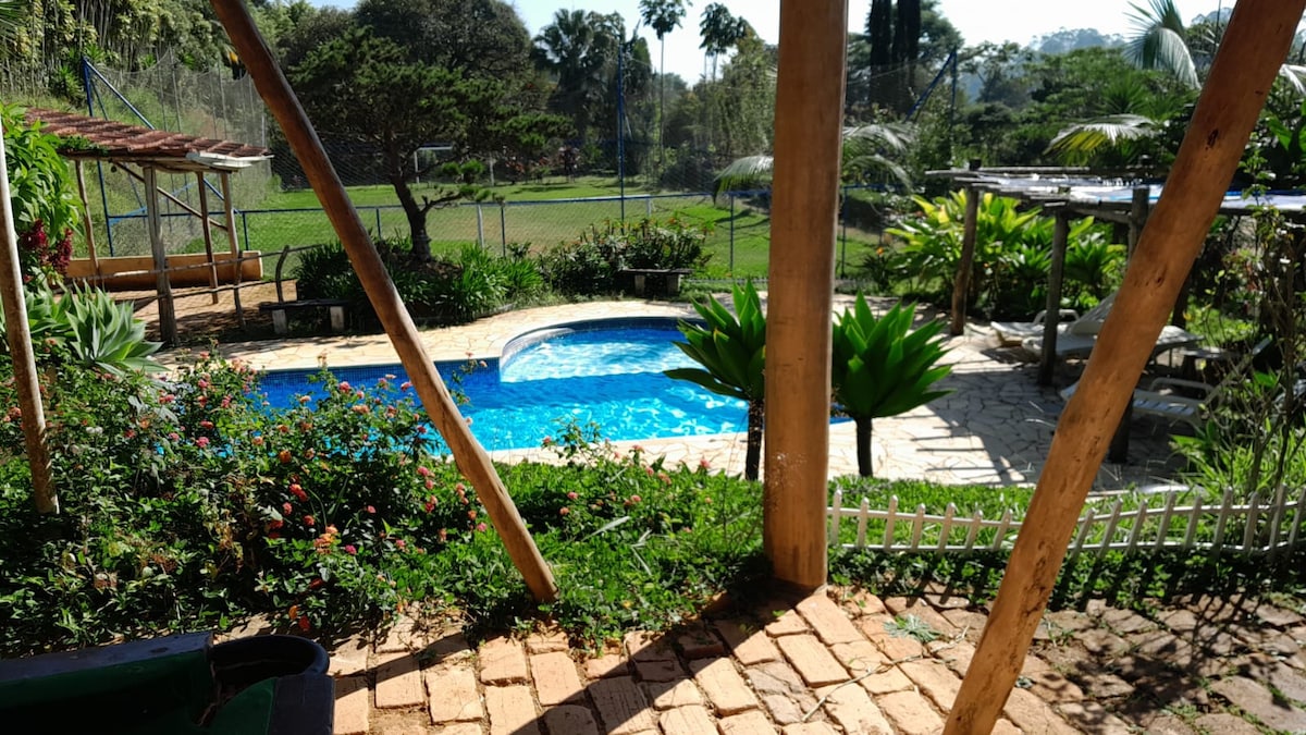 A scenic outdoor area is depicted, featuring a swimming pool surrounded by vibrant greenery and colorful flowers. The space is bordered by a white picket fence, with palm trees and a grassy field visible in the background.