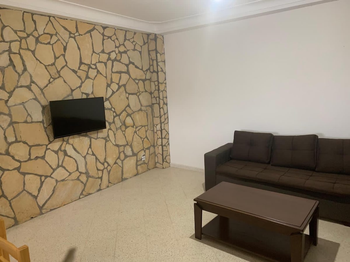 A living area is presented with a stone accent wall, featuring a mounted television. A dark sofa sits across from a wooden coffee table, creating a welcoming gathering space. The floor is smooth and light-colored, contributing to the open and airy ambiance.