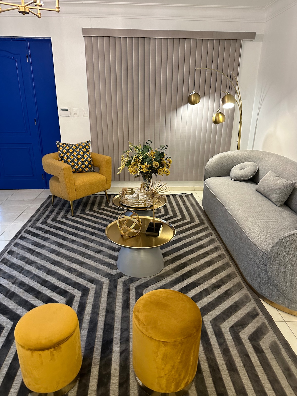 A modern living area features a light grey sofa and a yellow accent armchair with a patterned cushion. A round coffee table sits on a striped area rug, surrounded by two yellow stools. Soft natural light enters through vertical blinds, highlighting decorative plants.