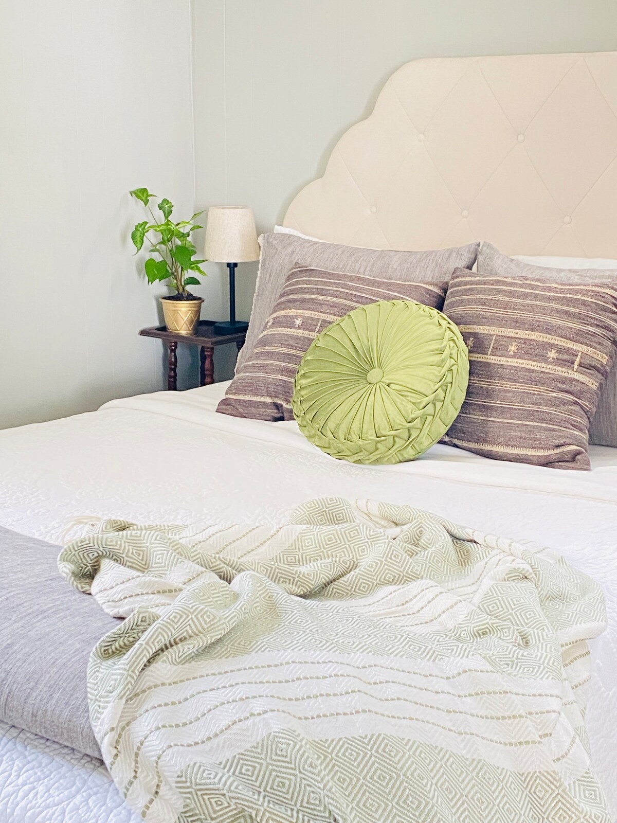 A cozy sleeping area features a spacious bed adorned with various decorative pillows, including a textured green accent. A small bedside table holds a potted plant and lamp, while a soft throw blanket is casually draped across the foot of the bed.