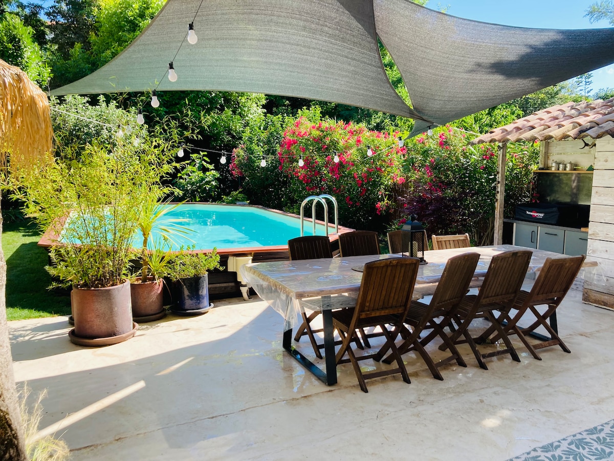 An outdoor dining area features a large wooden table with multiple chairs, shaded by a sail above. The nearby pool, surrounded by lush greenery and flowering plants, provides a relaxing backdrop. A barbecue station is visible in the corner, enhancing the inviting space.