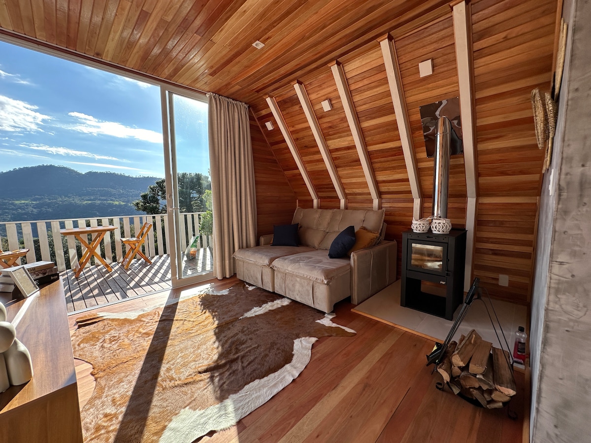 A spacious living area is illuminated by natural light from large glass doors. A retractable sofa is positioned near a modern wood-burning stove. A soft area rug complements the wooden flooring, while a small table and chairs are visible through the glass doors, offering outdoor access.