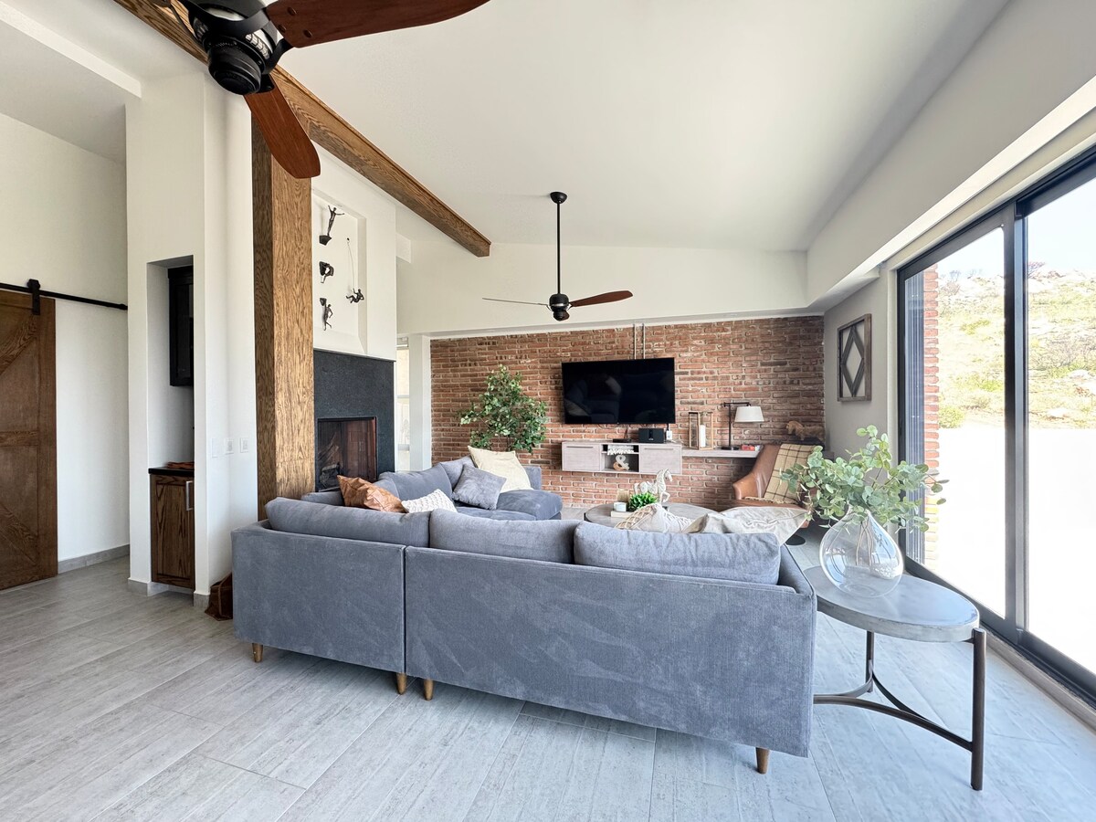 A modern living area features a comfortable grey sectional sofa, positioned near a wall-mounted television. Natural light fills the space through large glass windows, highlighting the exposed brick wall and the wooden ceiling beams. Decorative plants and a side table add a touch of warmth.