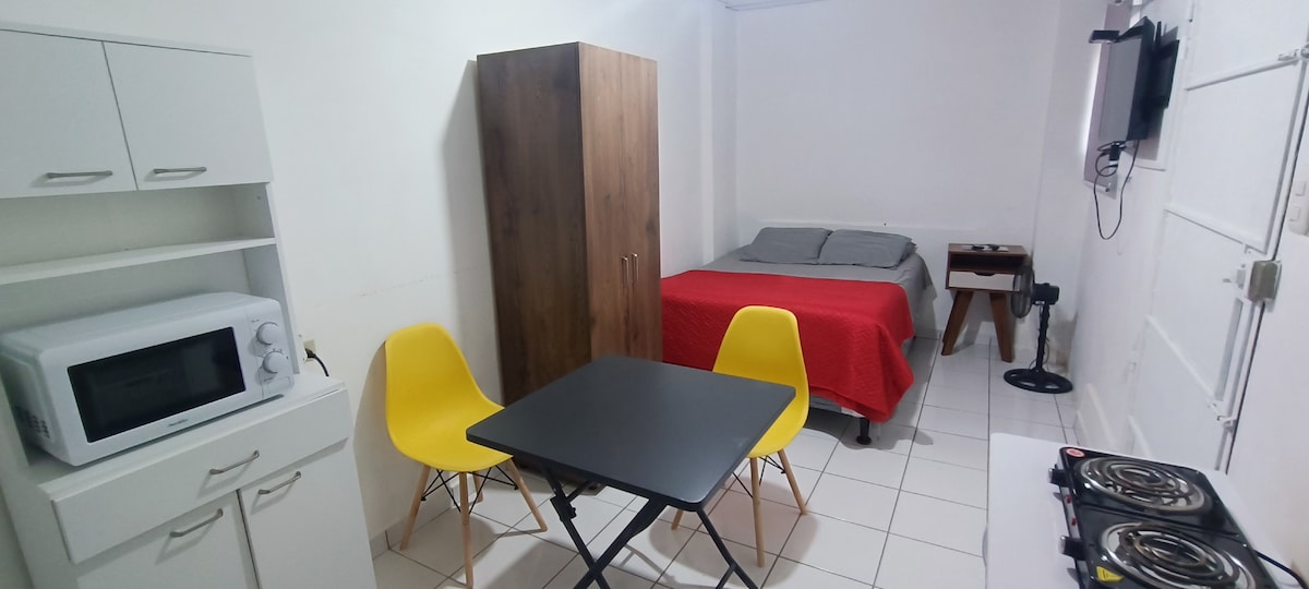 An efficient living space is shown, featuring a spacious bed covered with a grey blanket. A compact dining area with a black table and yellow chairs is present. A microwave and a wardrobe are situated in the corner, alongside a wall-mounted television.