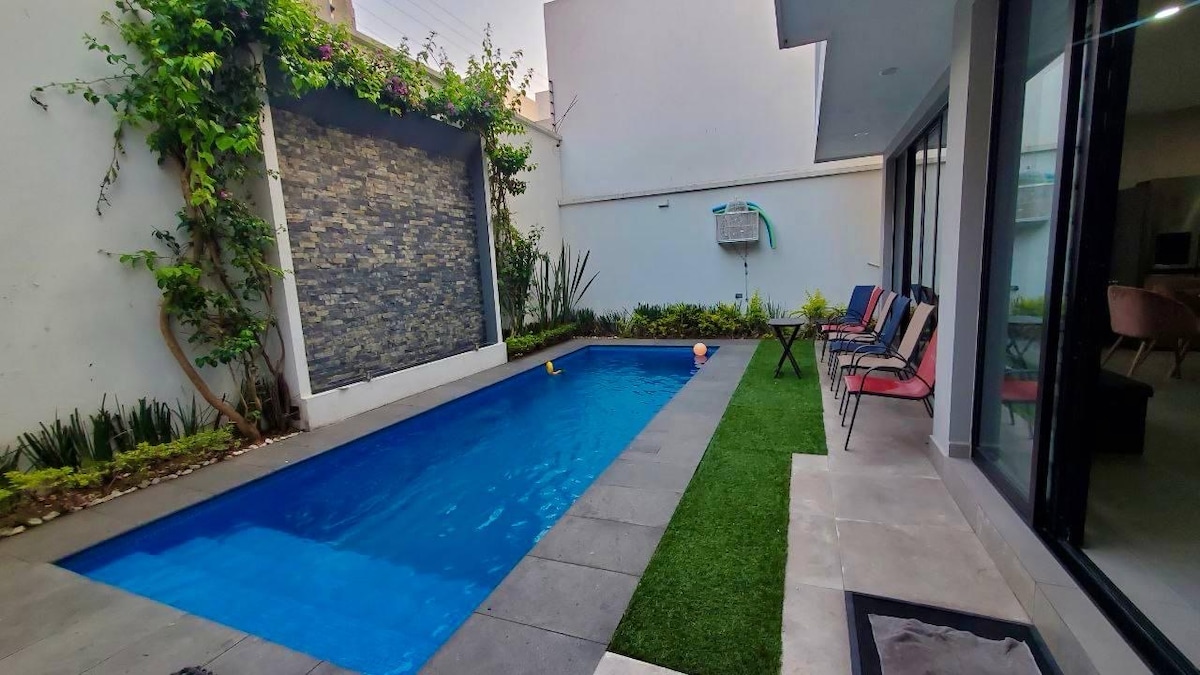 A serene outdoor area features a swimming pool surrounded by smooth stone tiles and a lush garden. Colorful chairs are arranged along one side, while a textured stone wall adds an element of interest. Sunlight reflects off the tranquil water, creating a calming setting.