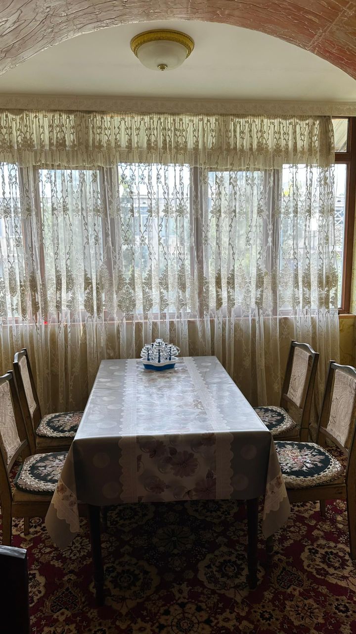A dining area is presented with a table covered by a light-colored tablecloth. Six wooden chairs are arranged around the table. Sheer lace curtains allow natural light to filter through, illuminating the space and providing a view of greenery outside.