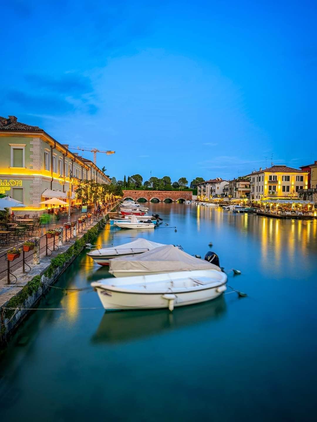 A serene waterfront scene features softly illuminated buildings lining the canal. Moored boats are seen on calm waters, reflecting the surrounding architecture. A bridge is visible in the background, framed by greenery, contributing to the tranquil atmosphere of the area.