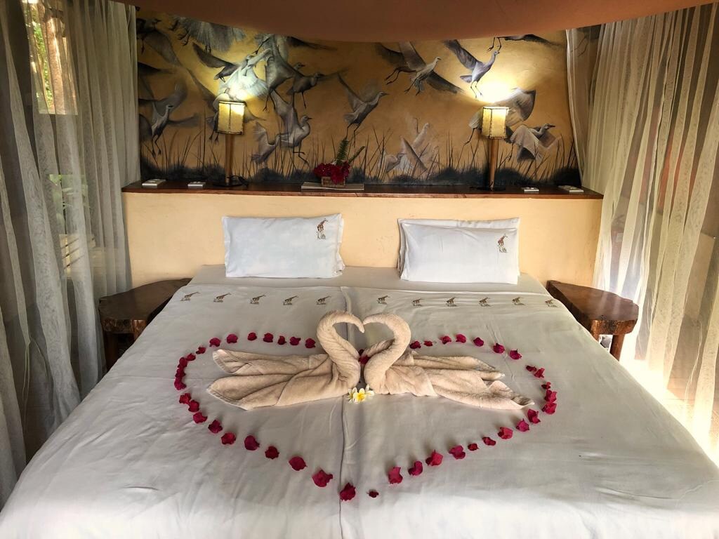 A spacious bed is adorned with a decorative arrangement of swan towels and rose petals, creating a heart shape on white linens. Soft, ambient lighting is provided by bedside lamps, while sheer curtains allow natural light to filter into the cozy room.