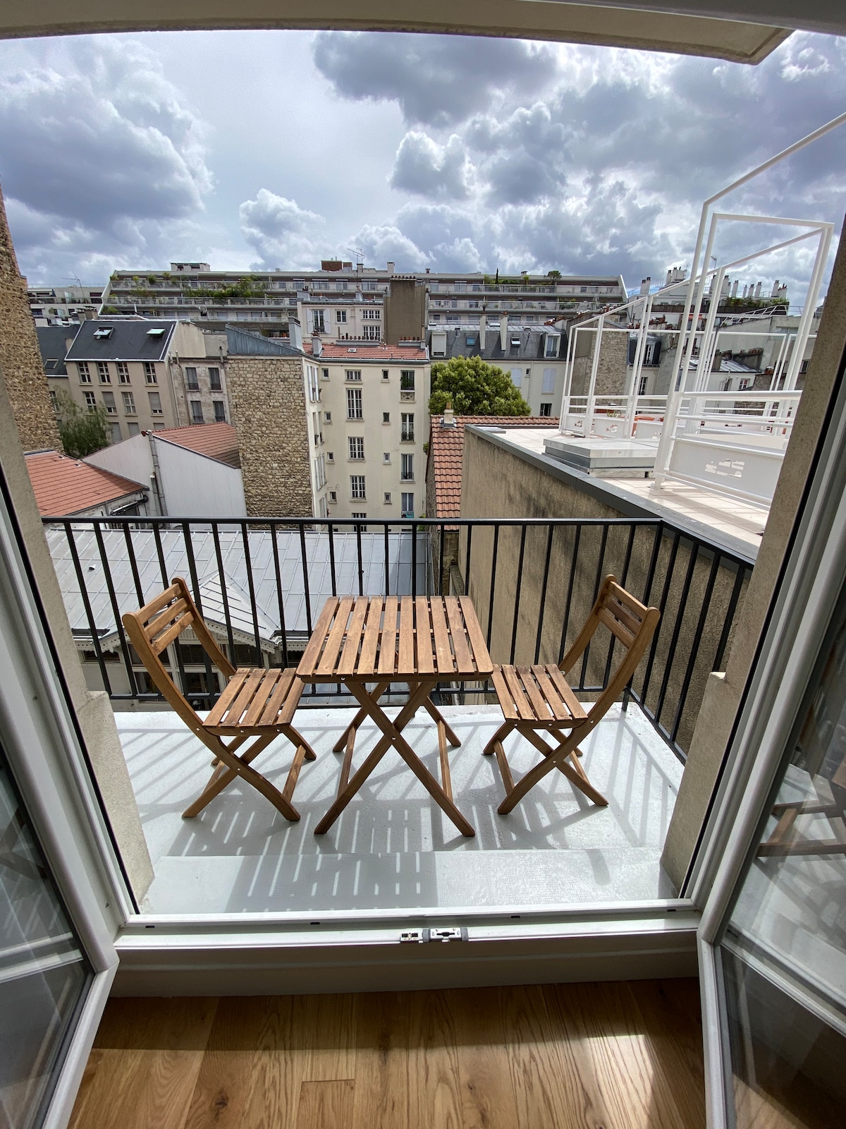 A small balcony features a wooden table with two folding chairs, offering a view of the surrounding rooftops and buildings. The balcony is framed by glass doors, allowing natural light to brighten the space.