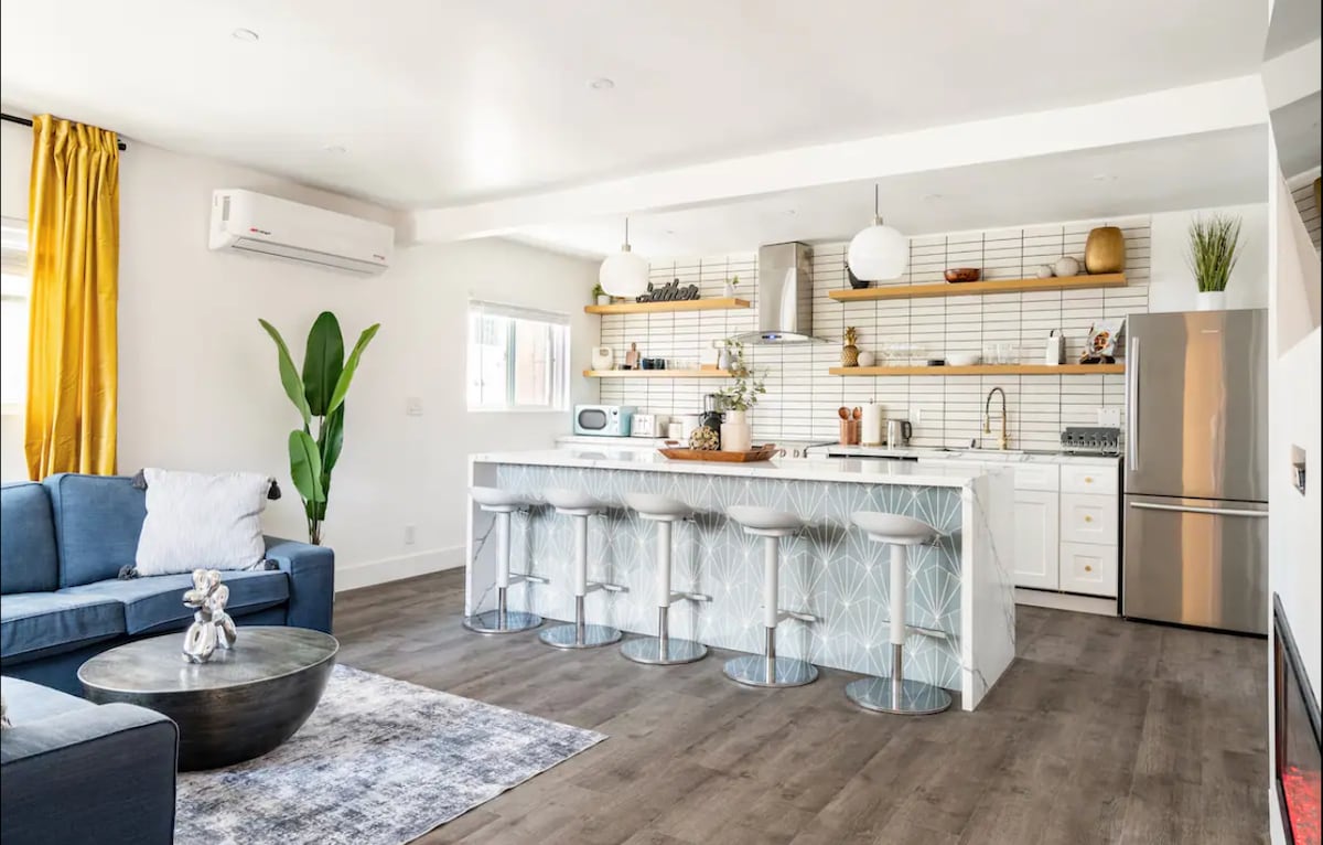 The modern kitchen features a spacious island with seating for four, complemented by stylish white cabinetry and a tiled backsplash. Natural light streams through a window, highlighting the contemporary appliances and decor. A cozy living area is situated nearby, adding to the inviting layout.