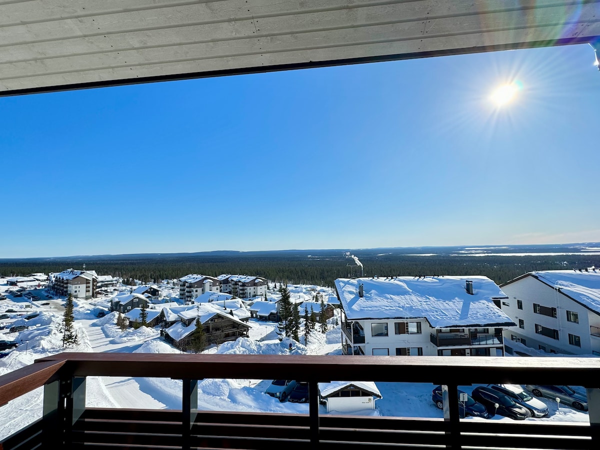 A panoramic view of a snow-covered landscape is presented from the balcony. The sky is clear and bright with sunlight shining down. Snow-draped roofs of nearby buildings are visible, along with a sprawling view of the forest in the distance.