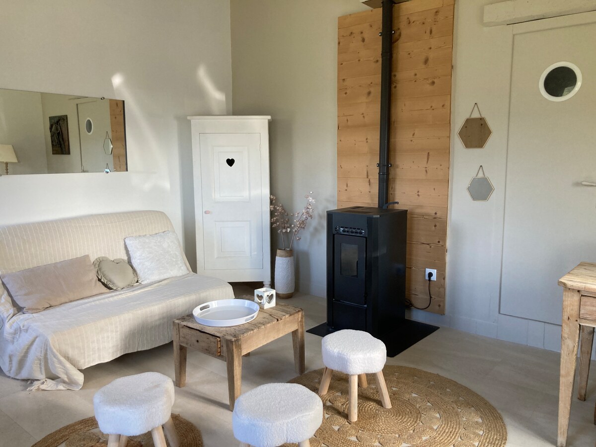 A cozy living area features a light-colored sofa accompanied by small, round stools. A rustic wooden table is placed in front, and a decorative rug adds warmth to the space. A pellet stove stands against the wall, providing a focal point in this inviting setting.