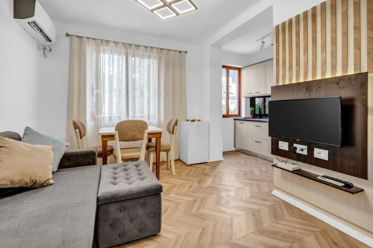 The living area features a gray sectional sofa with decorative cushions and a dining table for four situated near a window. A large, flat-screen television is mounted on a wooden wall panel. Bright natural light filters through sheer curtains, enhancing the inviting ambiance.