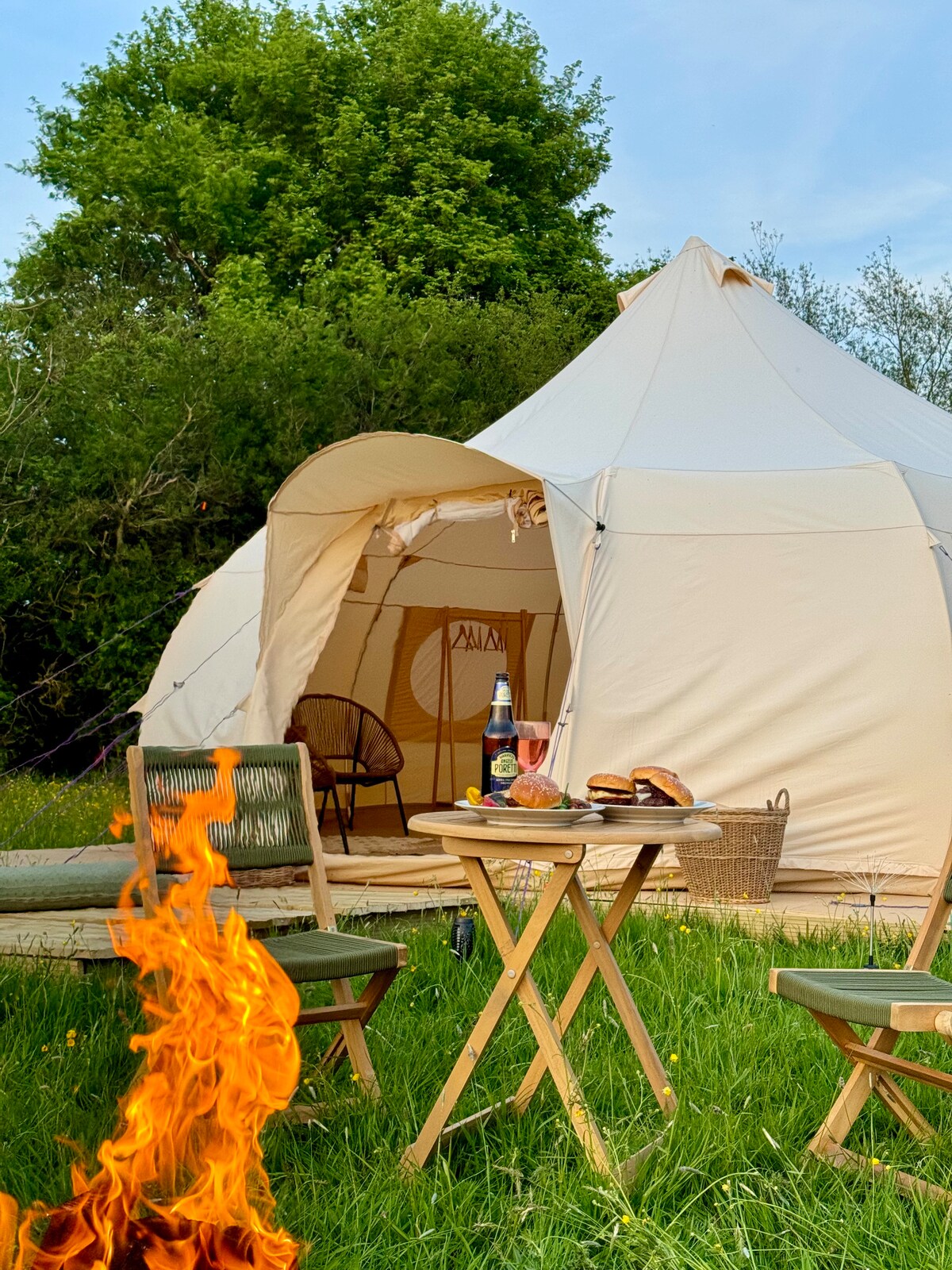 A spacious bell tent is featured under a bright sky, with an inviting entryway visible. A small fire burns in the foreground, with wooden tables set with plates of food and beverages nearby, surrounded by lush green grass.