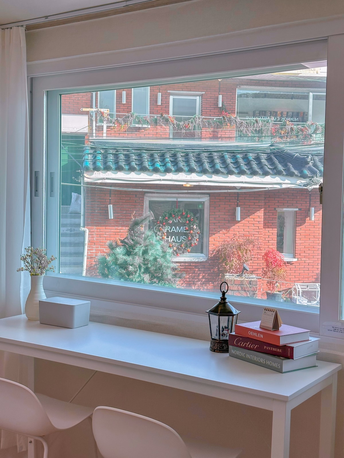 A view from a large window showcases neighboring buildings adorned with brick facades and lush greenery. A small table with decorative books and a lantern is positioned in front of the window, enhancing the inviting ambiance of the space.