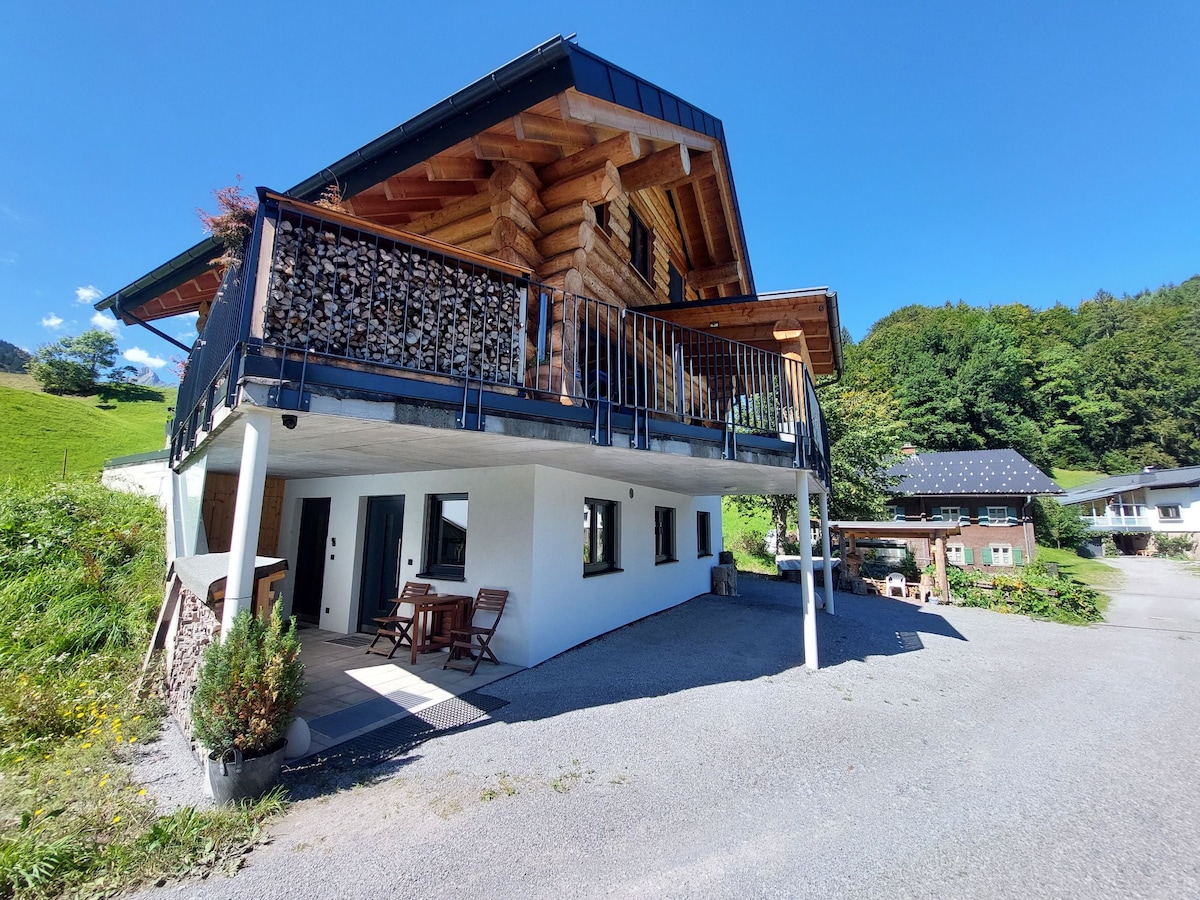 A wooden chalet is prominently featured, showcasing a sloped roof and a spacious balcony adorned with stacked firewood. Surrounding greenery is visible, along with a gravel pathway leading to the entrance. Nearby, additional residential buildings and solar panels can be seen in the background.