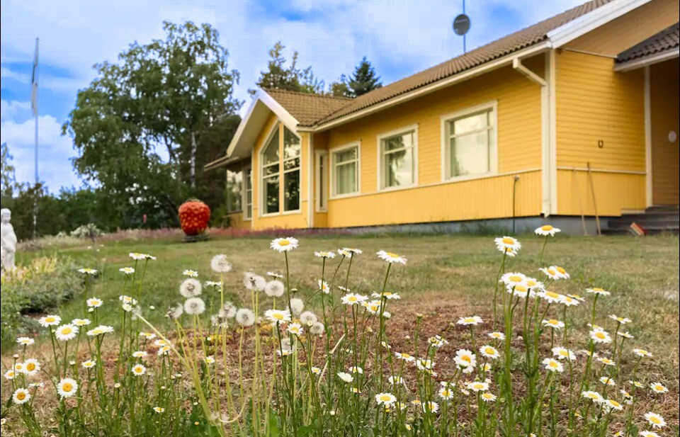 The yellow house is surrounded by a well-kept garden featuring vibrant flowers and a manicured lawn. A large window reflects natural light, while a garden sculpture adds a whimsical touch to the outdoor space. Trees provide a serene backdrop.