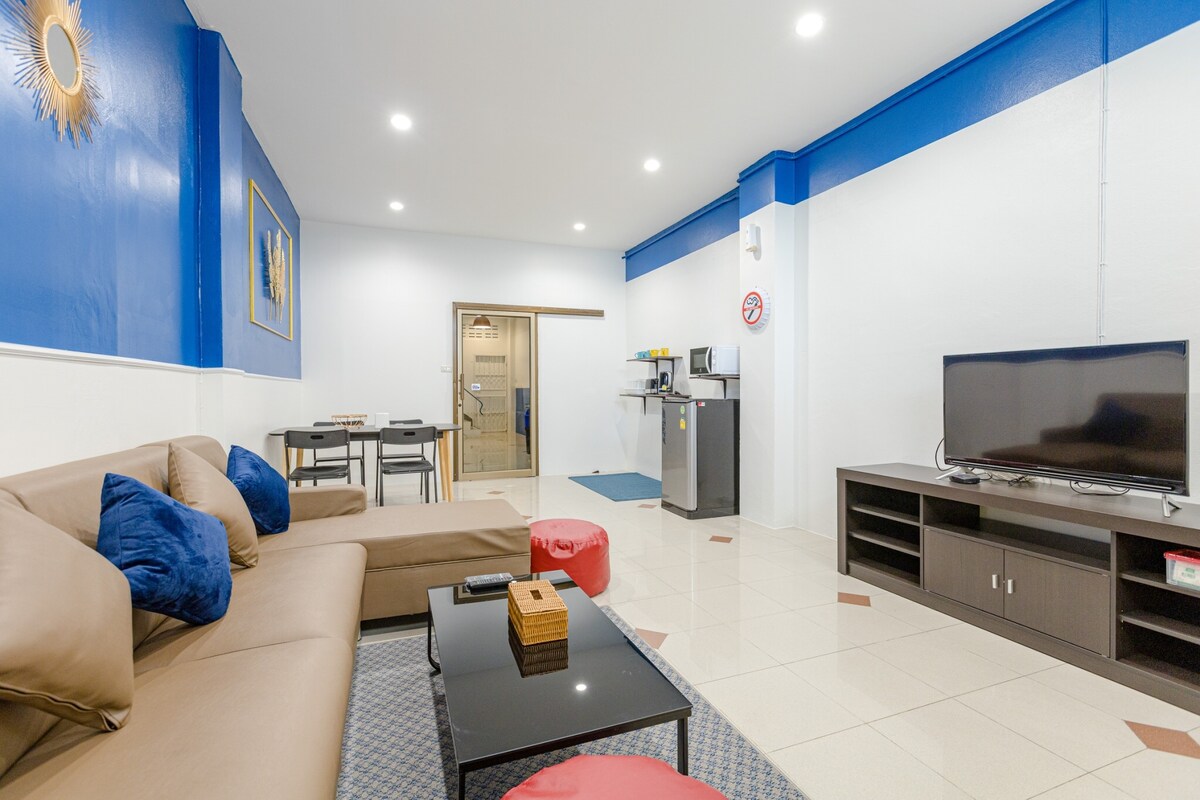A spacious living area features a comfortable sectional sofa adorned with colorful cushions. A flat-screen television is positioned on a dark media console. A dining table and chairs are present in the background, while a kitchenette is visible, equipped with essential appliances.