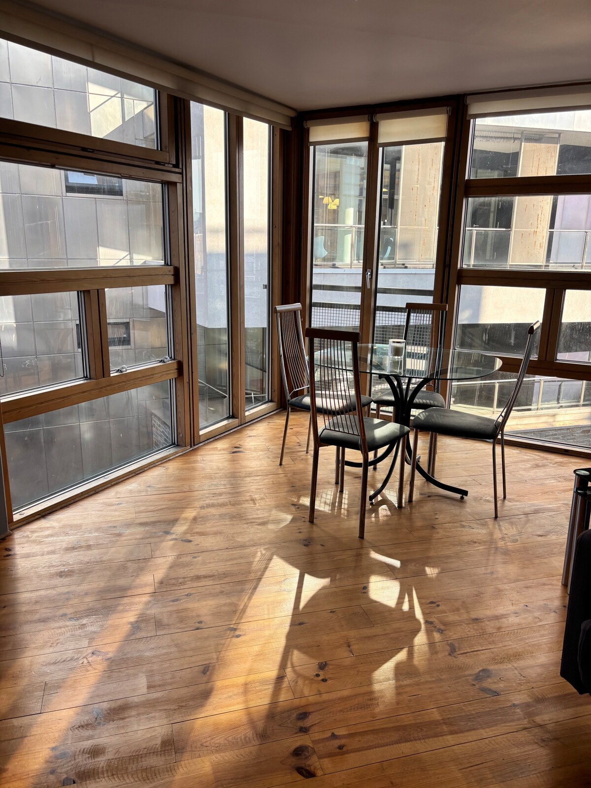 A dining area is illuminated by natural light, featuring a round glass table surrounded by four metal chairs. Floor-to-ceiling windows allow for ample sunlight and showcase a view of the neighboring buildings. The wooden floor adds a warm tone to the space.