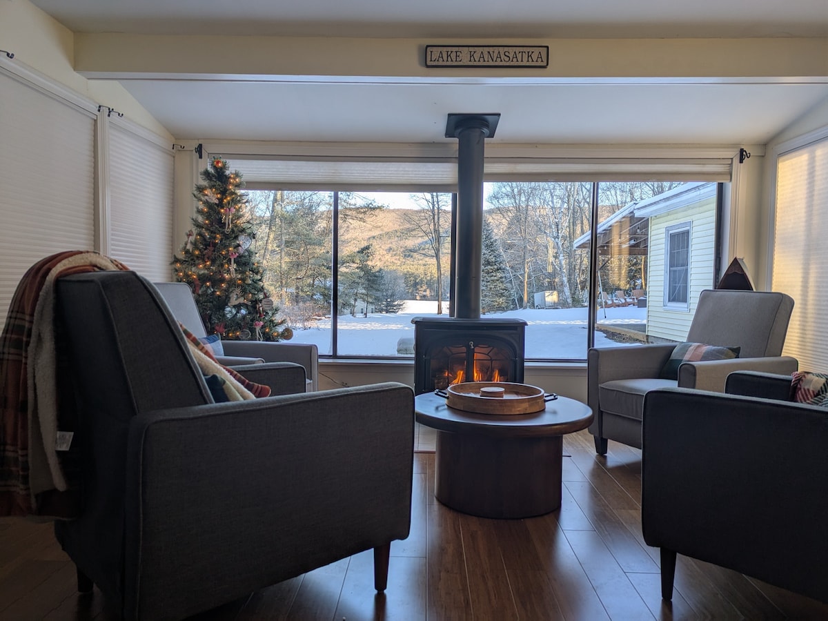 A cozy living area is highlighted by large windows showcasing the view of Lake Kanasatka and surrounding trees. Two armchairs face a circular coffee table in front of a gas stove, while a Christmas tree adds a festive touch to the space.