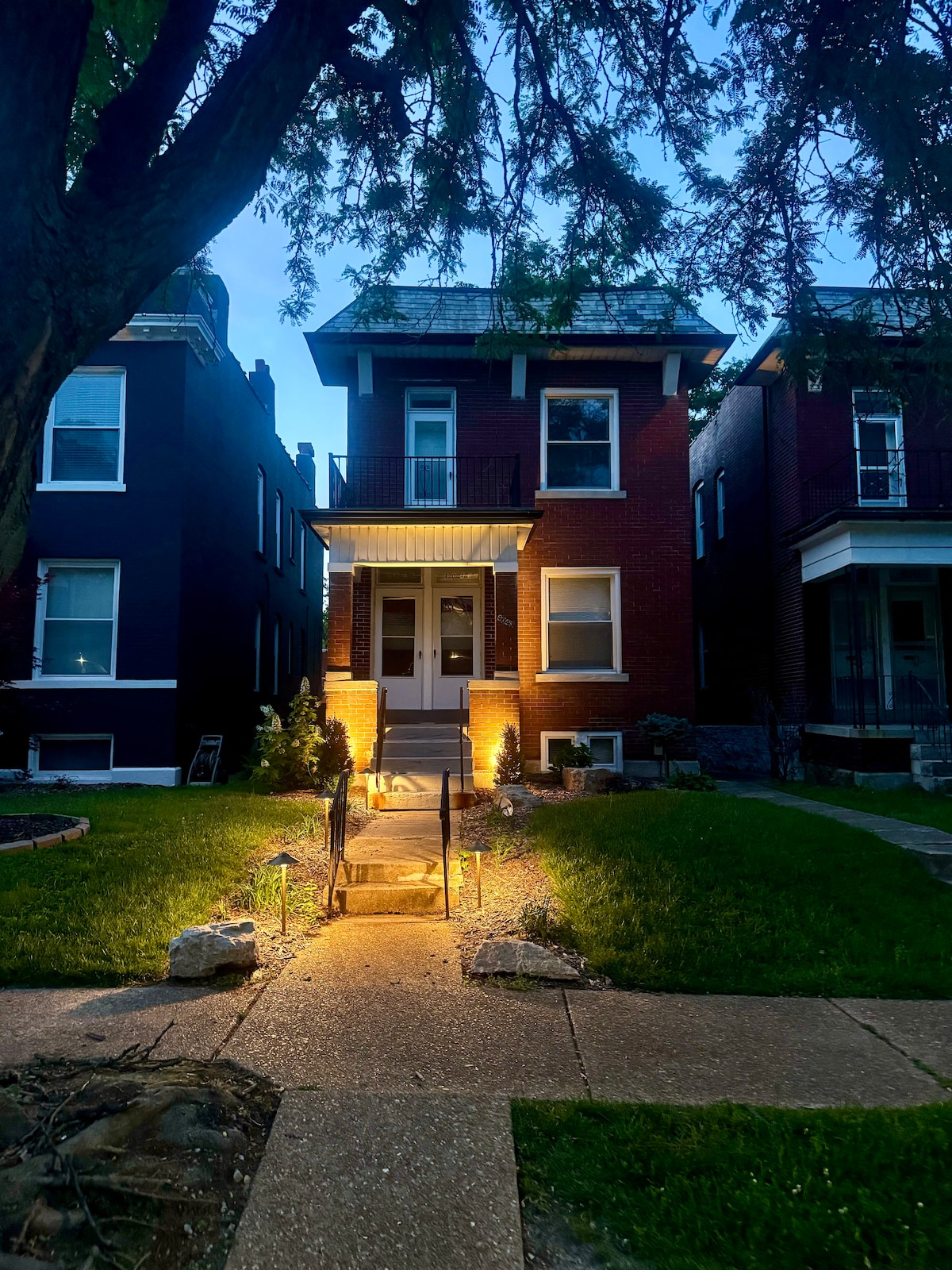 The historic duplex is illuminated in the evening light, highlighting its brick facade and inviting entrance. A pathway lined with stones leads up to the front steps, framed by lush green grass and bushes. Surrounding buildings are visible, showcasing a blend of architectural styles.