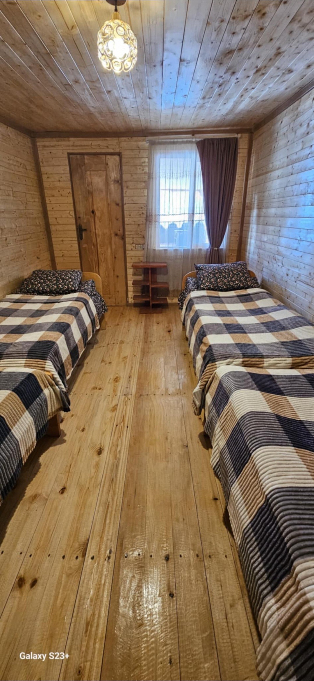 A well-lit room features two twin beds with plaid bedding, set against wooden walls and a polished wooden floor. A window dressed with a dark curtain allows natural light to enter. A small shelf is positioned near the door, adding to the functional space.