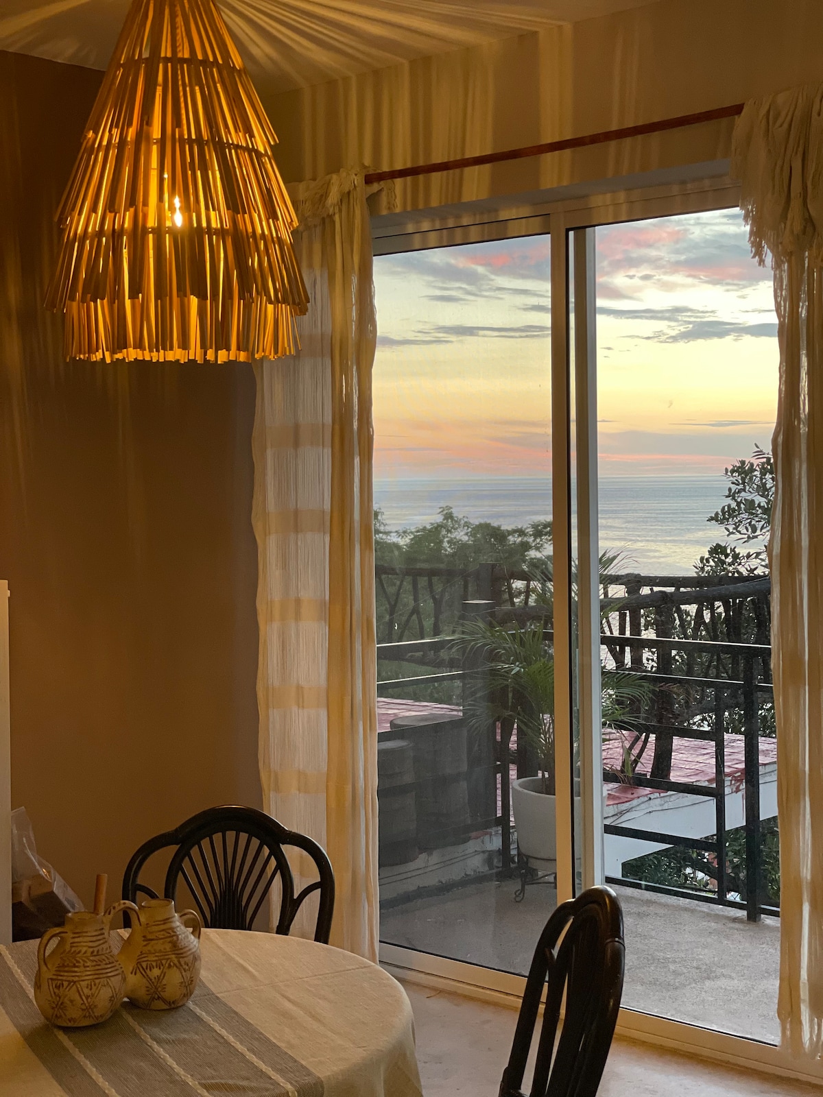 A dining area is illuminated by a unique woven pendant light, creating a warm ambiance. Large glass doors provide expansive views of the ocean and sunset, inviting natural light into the space. A white tablecloth covers the round table, accompanied by two decorative ceramic vases.