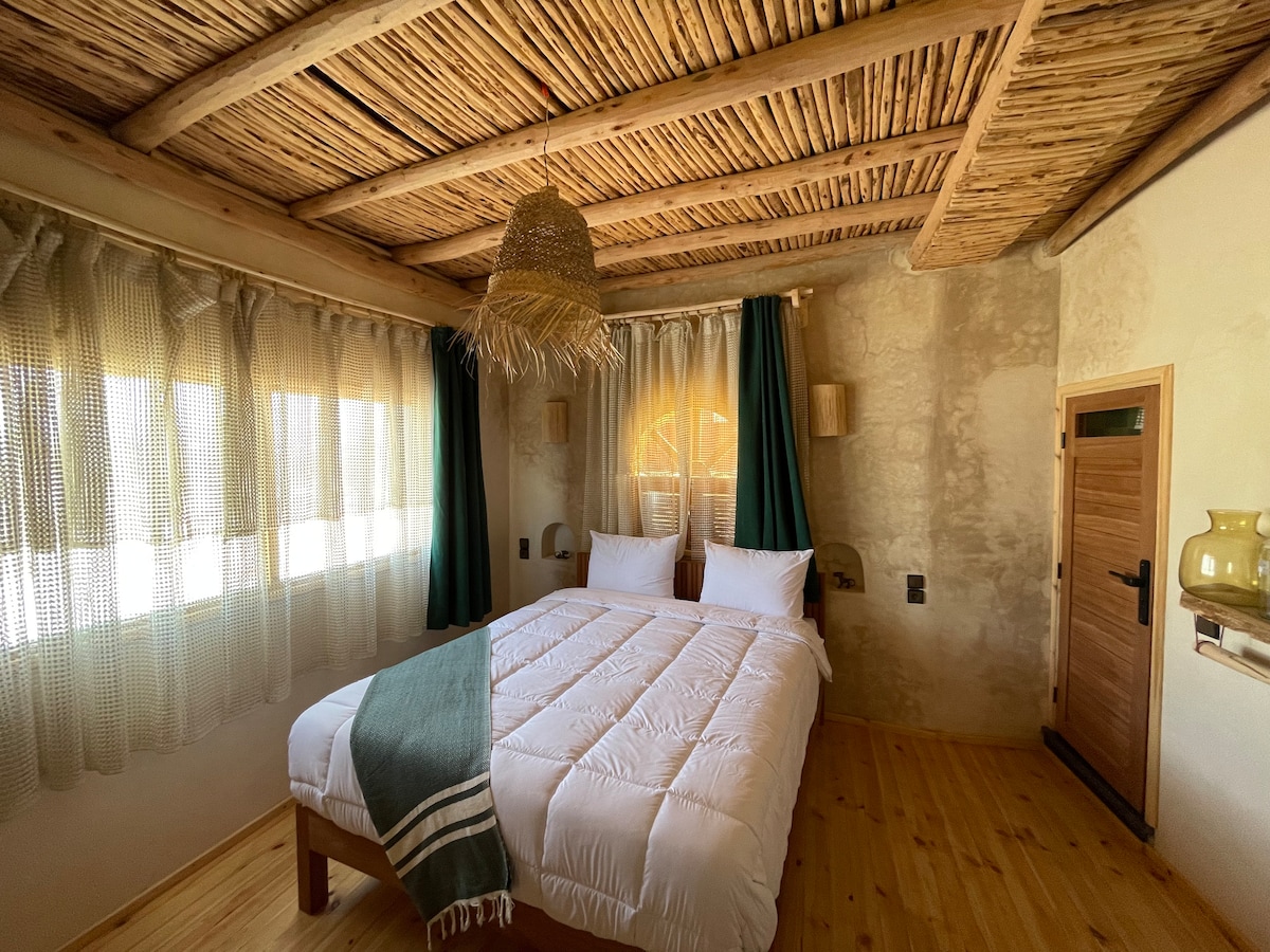 A cozy bedroom features a spacious bed with crisp white bedding. Natural wood beams adorn the ceiling, while green curtains frame the windows, allowing filtered light to enter. Textured walls add warmth, and a woven light fixture hangs above, contributing to the tranquil atmosphere.