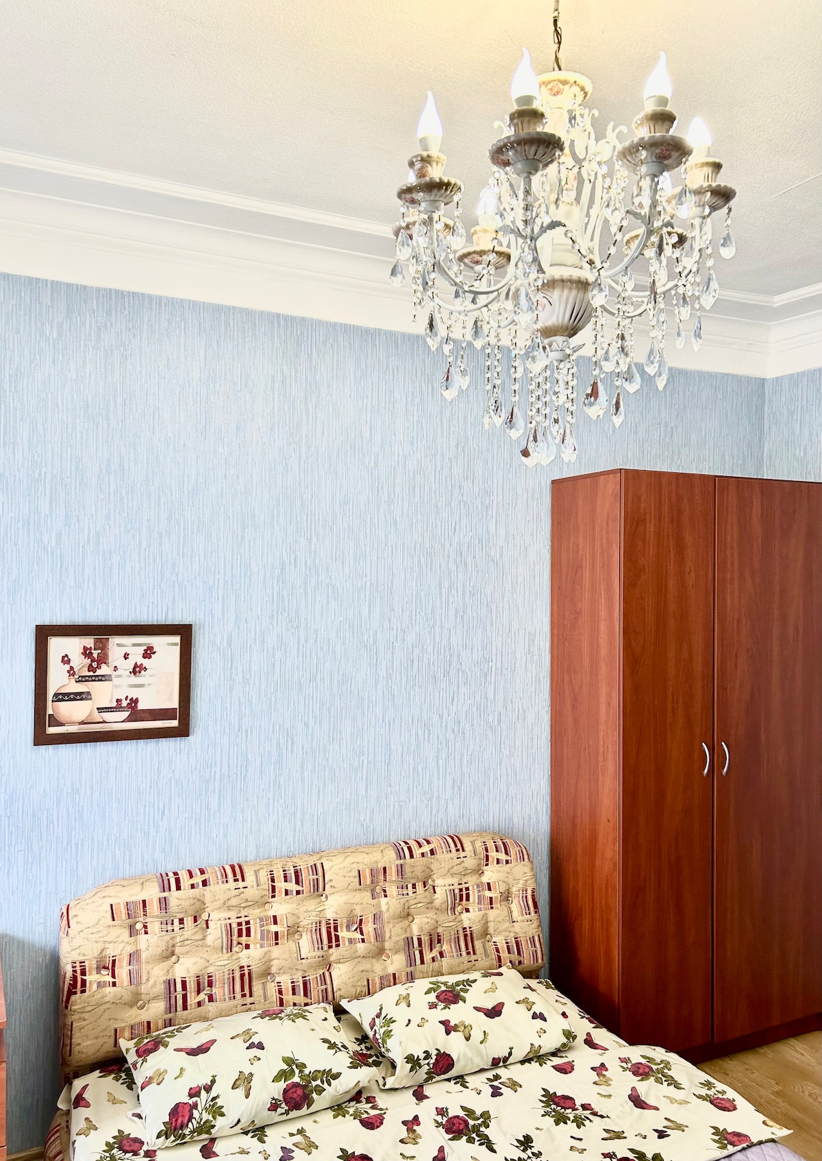 A cozy bedroom is showcased with a floral-patterned bedspread draped over a spacious bed. A wooden wardrobe with two doors stands nearby. A crystal chandelier hangs from the ceiling, providing soft illumination, and a framed picture featuring a beverage theme is mounted on the wall.