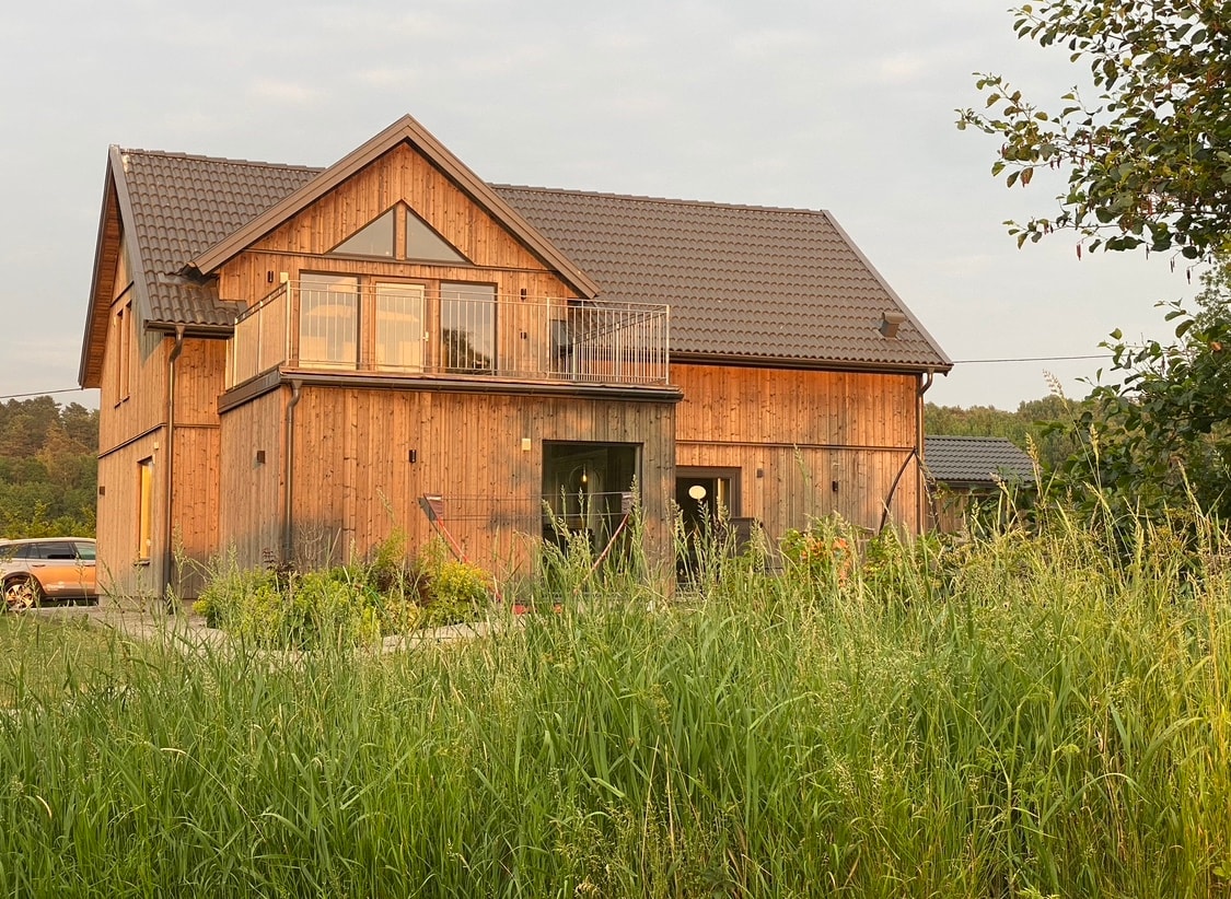 A two-story wooden house is seen among tall grasses, showcasing a mix of natural tones. The upper level features a balcony with railing, while large windows illuminate the lower space. A driveway with parked vehicles is visible in the foreground, complementing the serene rural setting.