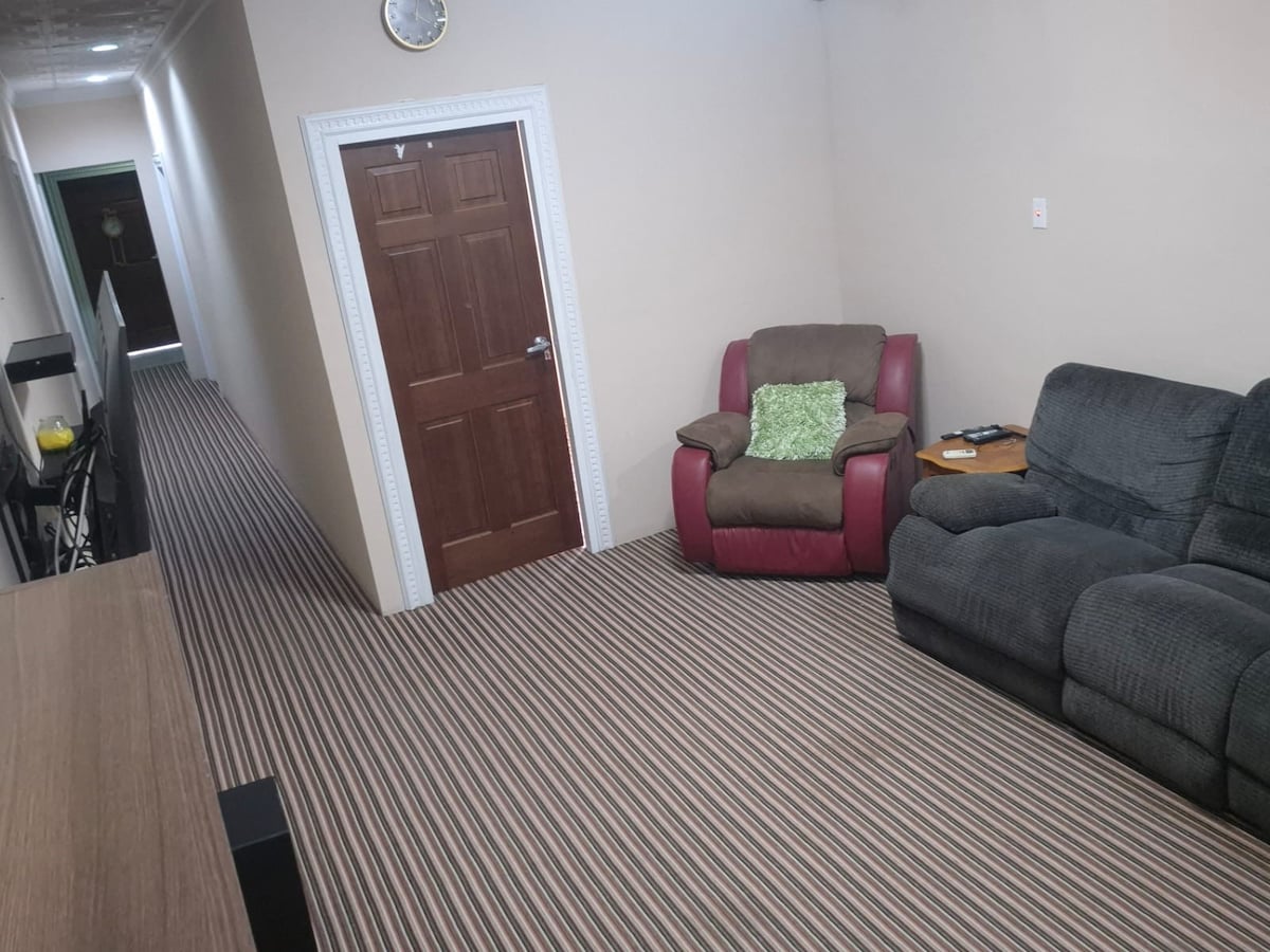 A hallway is shown with striped carpet leading toward a closed wooden door. Two comfortable seating options are present: a grey sofa and a brown armchair with a green pillow. A small table with a lamp sits beside the armchair, contributing to the cozy atmosphere.