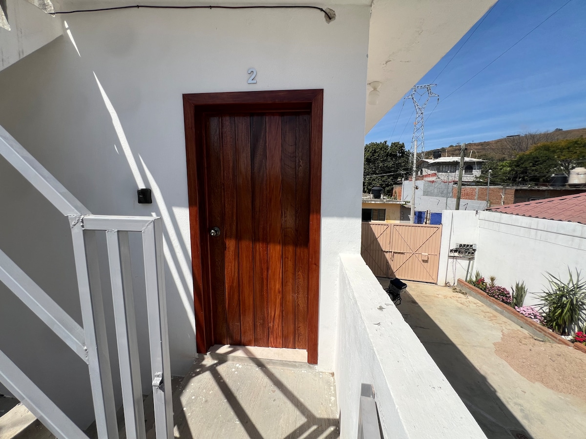 A wooden door numbered '2' is positioned at the end of a clean balcony. Sturdy white railings frame the space, while a clear blue sky and surrounding structures are visible in the background.