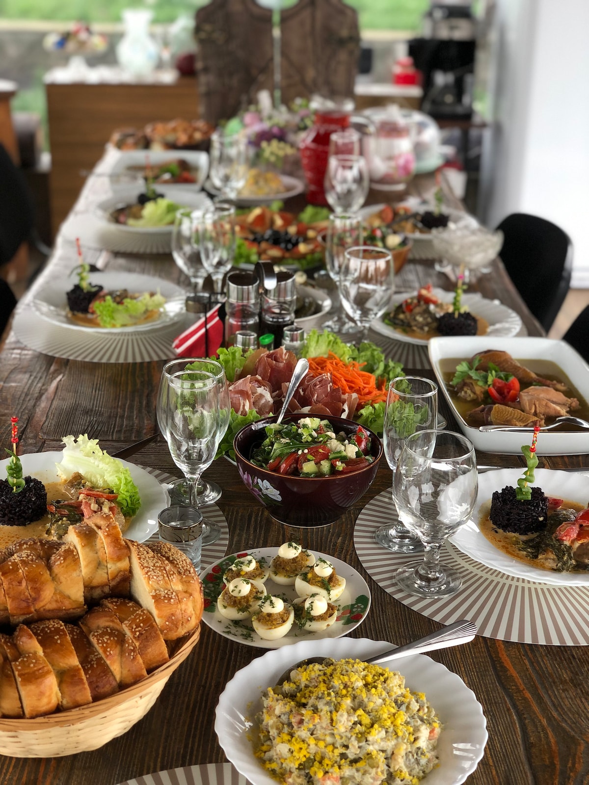 A dining table is set with a variety of dishes, featuring salads, sliced meats, and colorful vegetables. Glassware is arranged neatly alongside the plates. Freshly baked bread is displayed on one end, while garnishes add detail to the meal presentation.