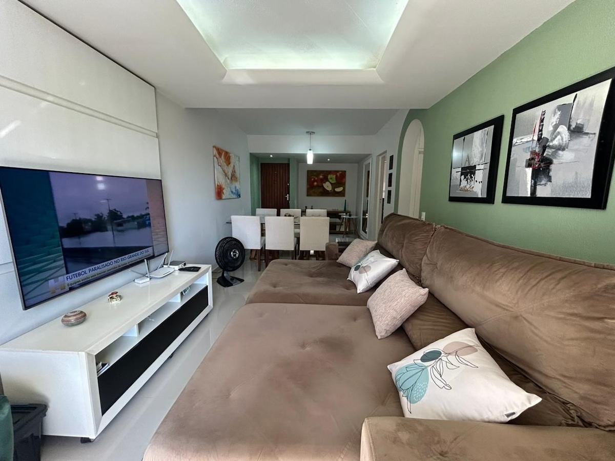A spacious living area features a large, retractable sofa adorned with decorative pillows. A flat-screen Smart TV is mounted on the wall opposite. Light green walls complement framed artworks, creating an inviting ambience. The open layout transitions into the dining area with a table and chairs.