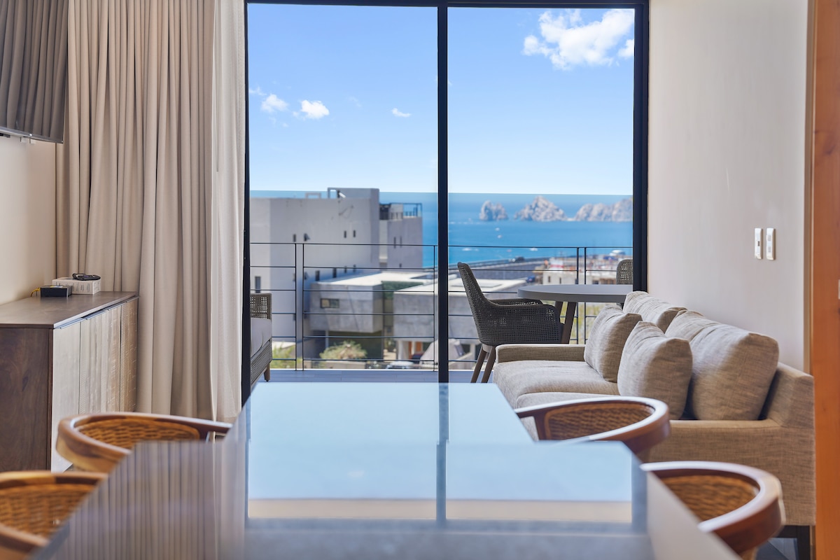 A modern living area features a spacious sofa arranged next to a large glass window, offering views of the ocean and the famous Arch in the distance. A dining table is positioned in the center, with natural light illuminating the space.