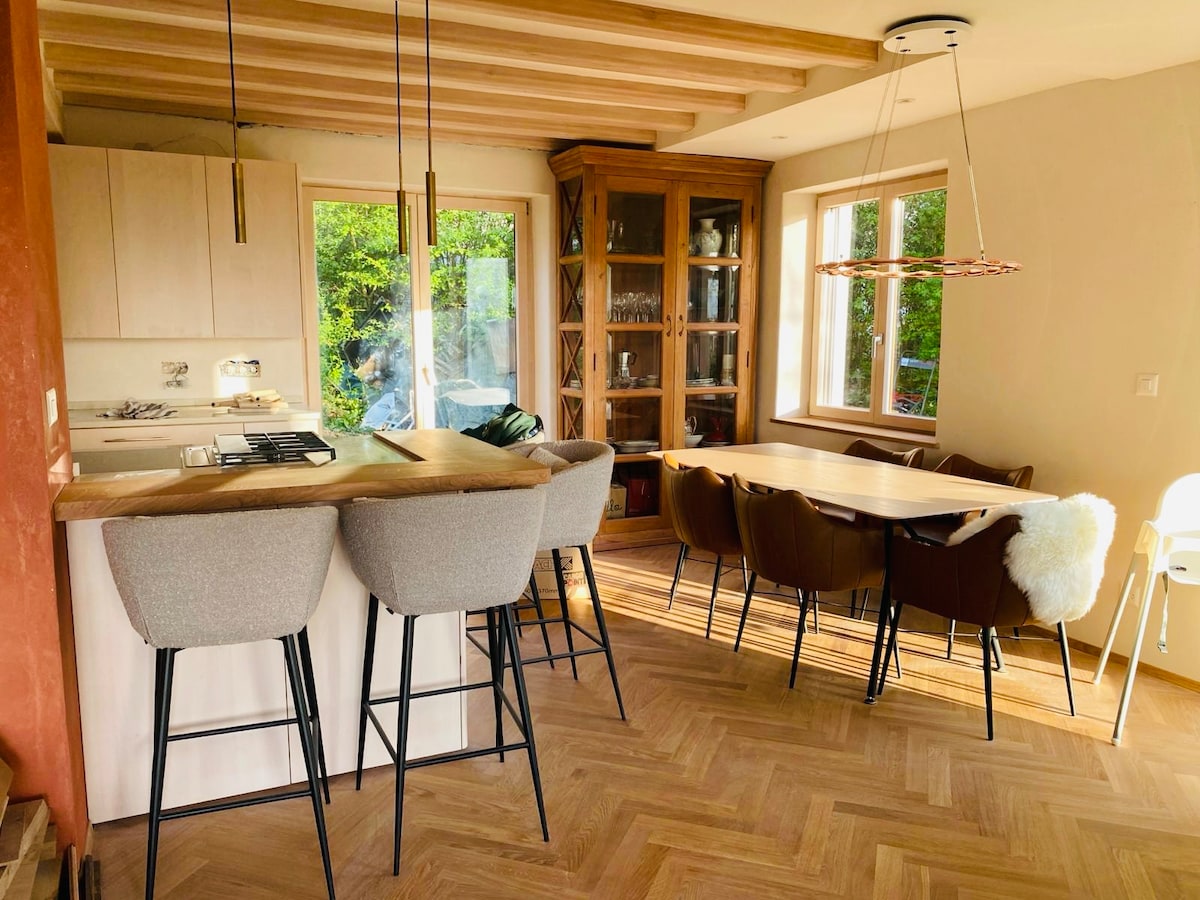 A bright dining area features a wooden table surrounded by various chairs, including upholstered options. A kitchen island with bar stools is situated nearby. Large windows allow natural light to fill the space, showcasing views of the greenery outside.