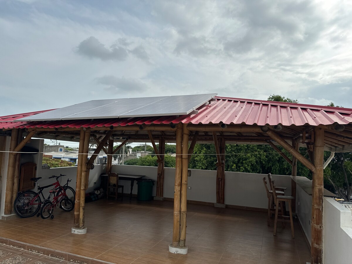 An outdoor space features a red-roofed gazebo constructed from bamboo. Solar panels are positioned atop the structure, providing shade. A tiled floor offers a clean surface, while two bicycles are visible in one corner, accompanied by a green trash bin and a wooden chair.