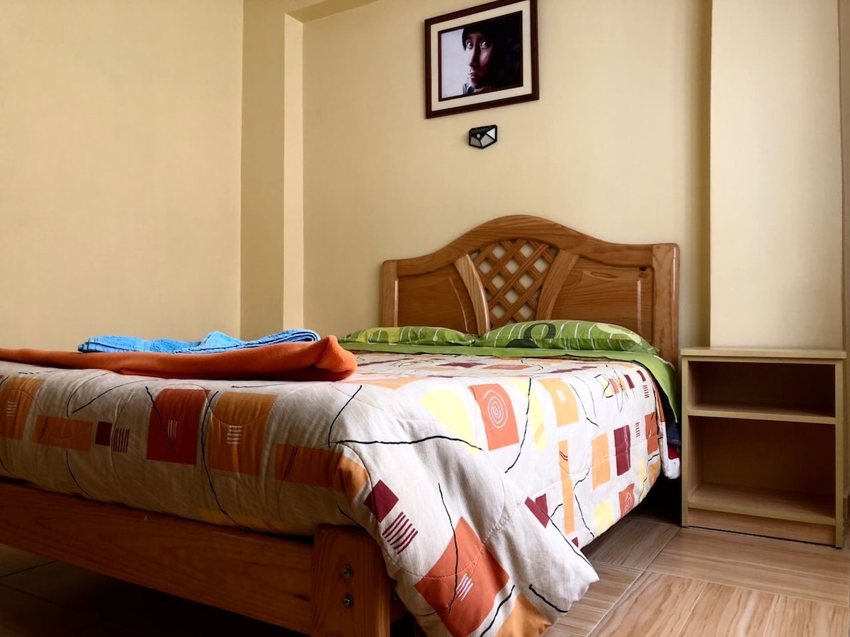 A cozy sleeping area is presented with a wooden bed featuring a decorative headboard and colorful bedding. A nightstand is positioned beside the bed, and a framed picture is mounted on the wall above. Natural light is filtered through the room, enhancing the warm tones.