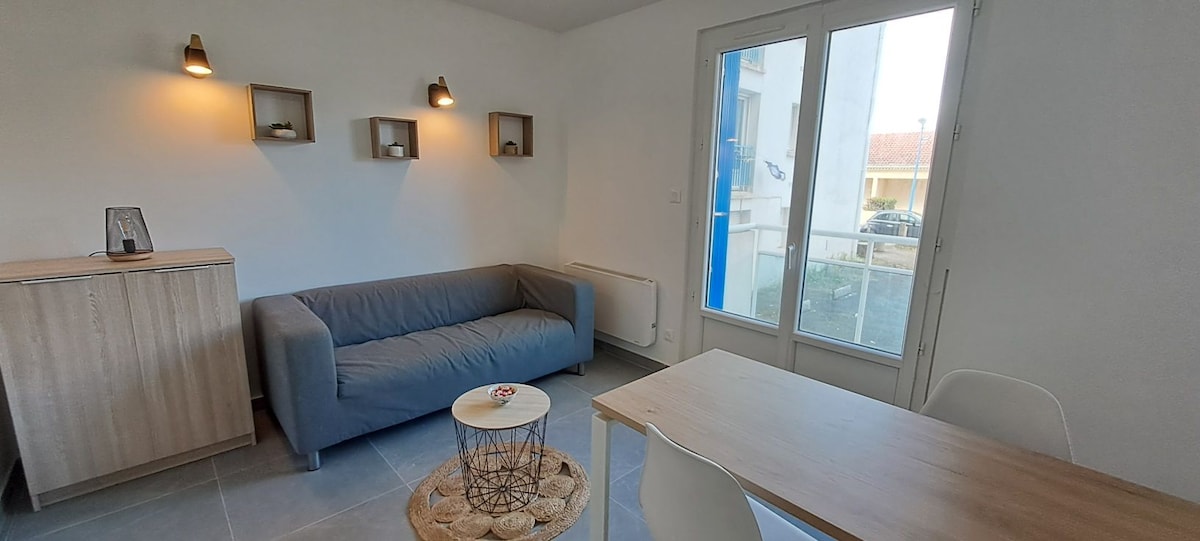 A well-lit living area is presented with a gray sofa across from a wooden cabinet with decorative shelves. A round table made of natural materials sits beside the window, where light enters through large glass doors that lead to a small outdoor space.