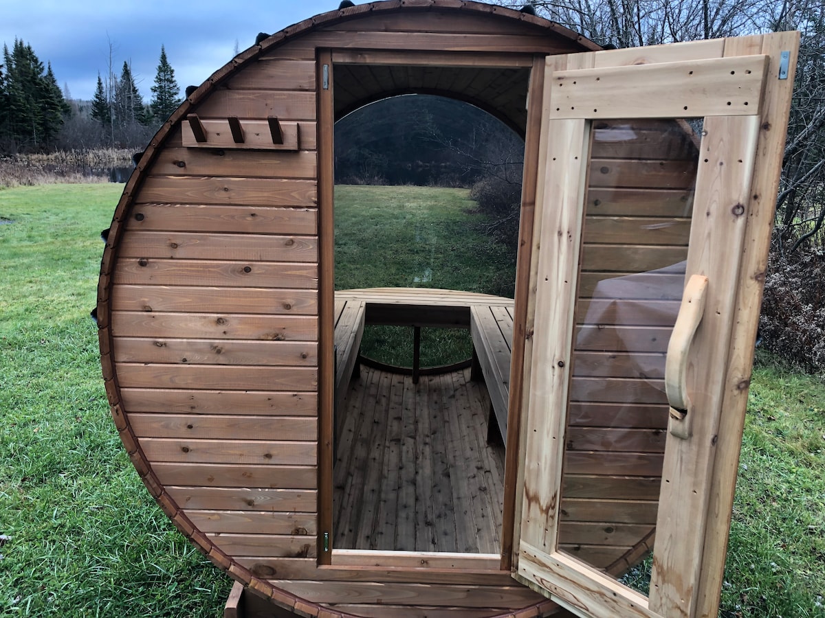 A wooden sauna structure with an oval shape stands amidst green grass. The double door is open, revealing a simple interior with wooden benches along the sides. Natural surroundings enhance the peaceful setting, and trees are visible in the background under a cloudy sky.