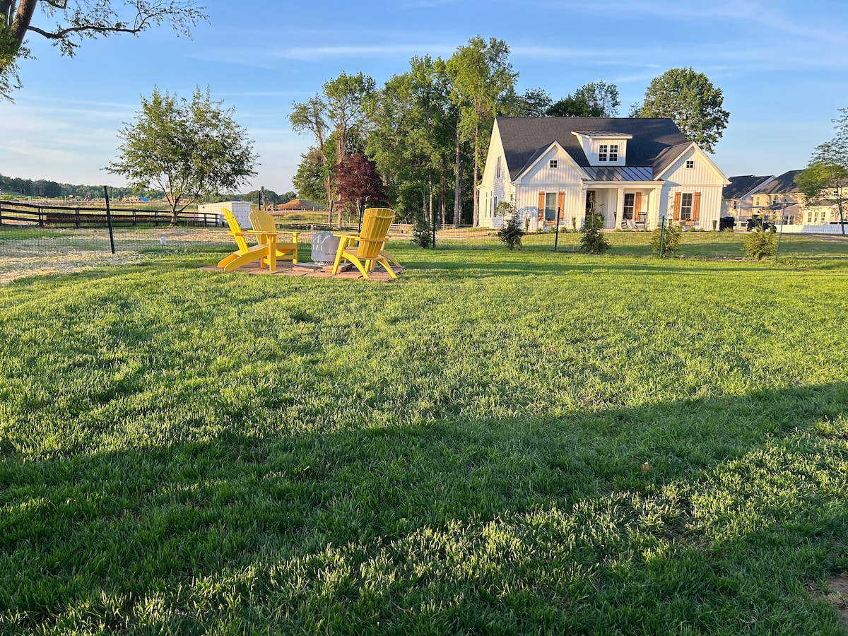 A spacious yard features vibrant green grass and two bright yellow adirondack chairs positioned around a small table, perfect for enjoying the outdoors. The charming cottage is viewed in the background, surrounded by trees and a fenced area.