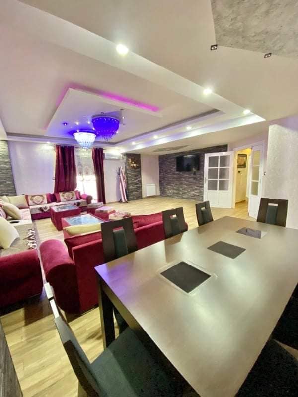 A dining area is visible in the foreground, featuring a large table with built-in outlets. The spacious living room showcases a mix of seating options in rich red hues, adorned with decorative cushions. Ambient lighting is provided by a chandelier, enhancing the overall space.