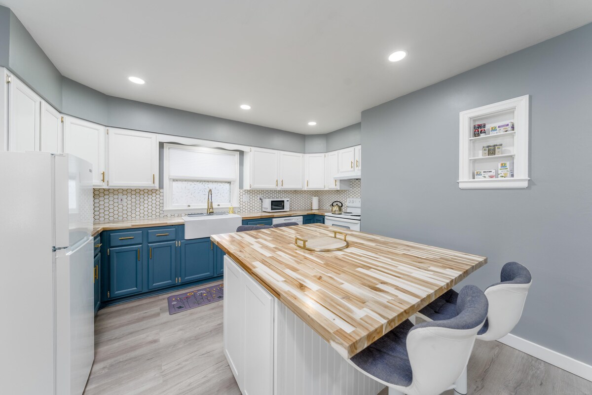 A modern kitchen features blue lower cabinets and white upper cabinets, complemented by a wooden countertop island. Bright lighting illuminates the space, showcasing stainless steel appliances and a small shelf with decor. An inviting seating area with two upholstered chairs is positioned around the island.