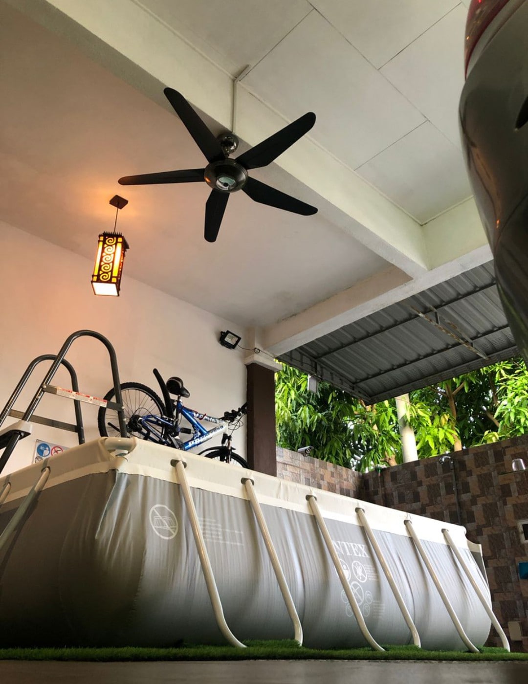 An outdoor area features a rectangular above-ground pool, surrounded by a well-manicured lawn. A ceiling fan is positioned above, providing cooling air. A warm light fixture hangs overhead, adding a cozy touch to the space, which also accommodates a bicycle on a rack.