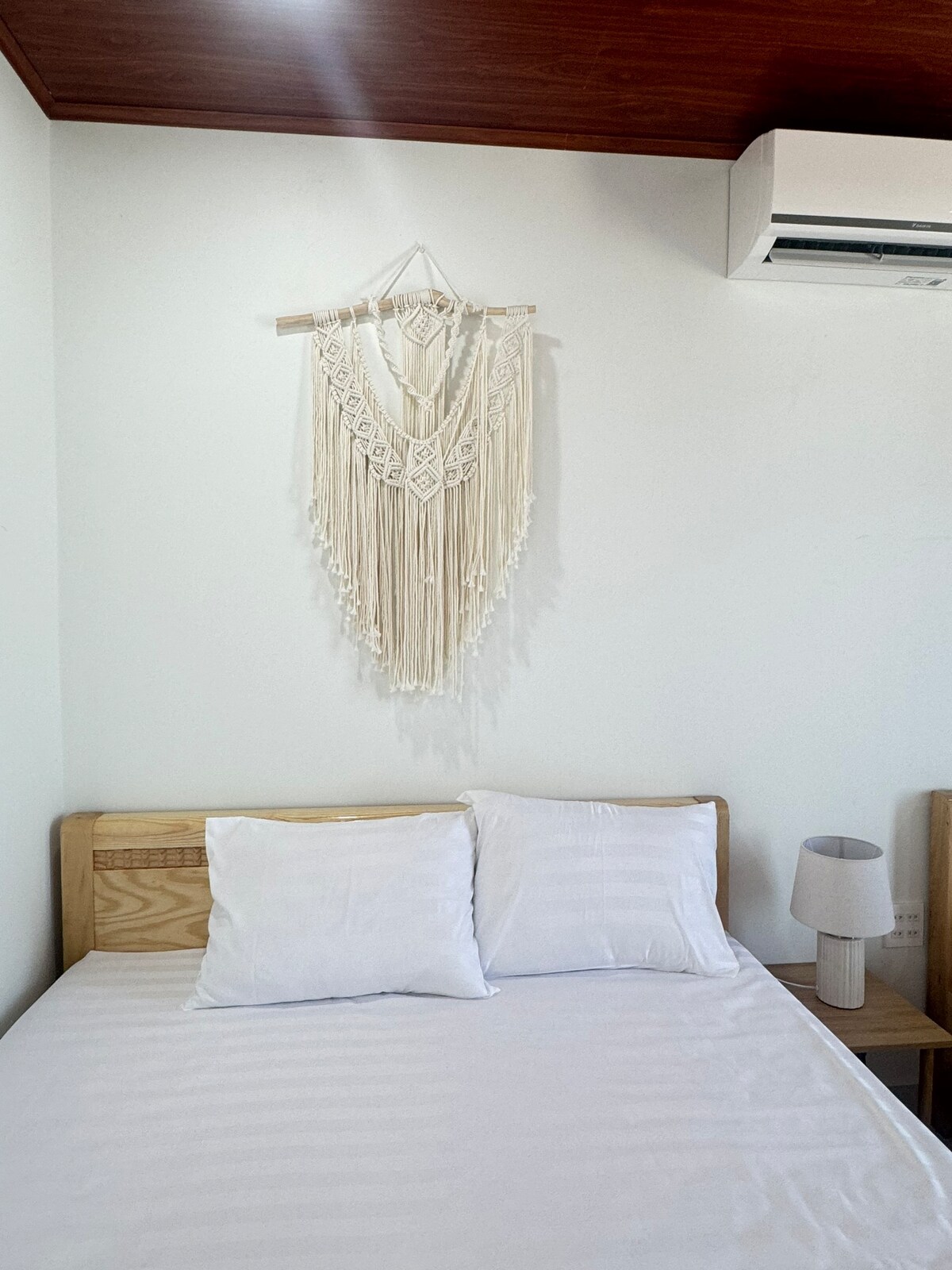 A sleeping area features a spacious bed with two white pillows, complemented by wooden nightstands. A decorative macramé wall hanging adds texture above the bed, while a small lamp sits on one nightstand. An air conditioning unit is mounted on the wall.