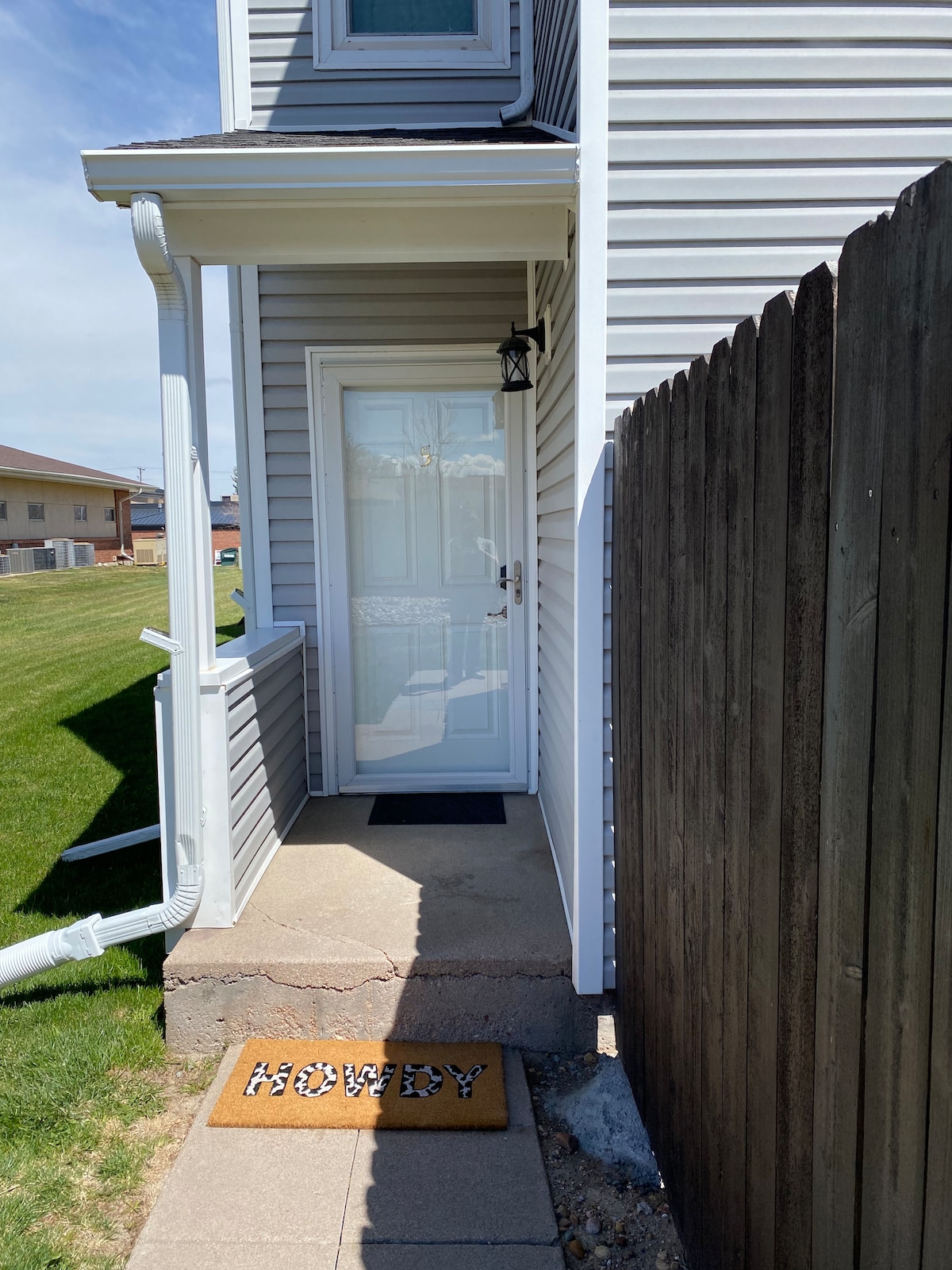 The entrance to the condo is shown, featuring a clean, white door framed by a small porch. A 'HOWDY' doormat is positioned on the stone pathway leading to the entrance. Grass lines the side of the walkway, providing an inviting outdoor space.