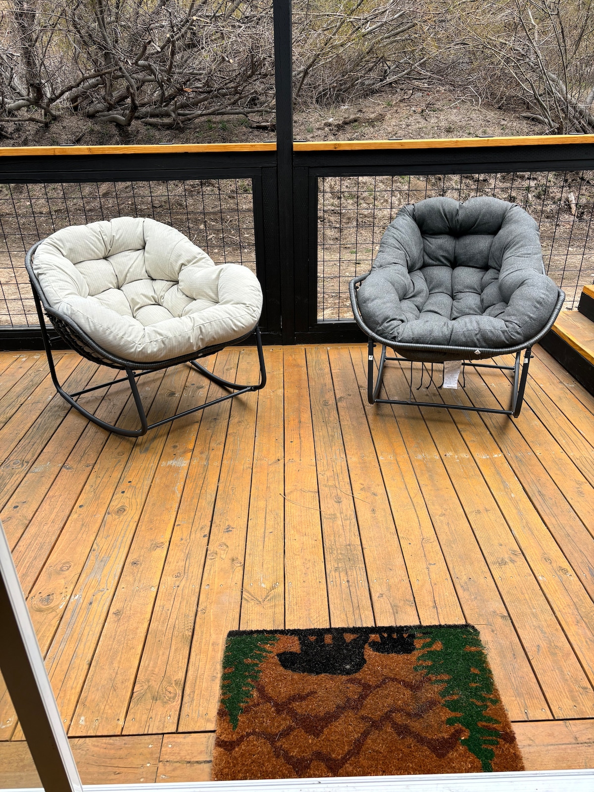 A cozy sunroom features two cushioned chairs in neutral tones, providing comfortable seating for relaxation. The wooden deck is partially shaded, surrounded by natural scenery. A bear-themed welcome mat adds a touch of charm at the entrance.